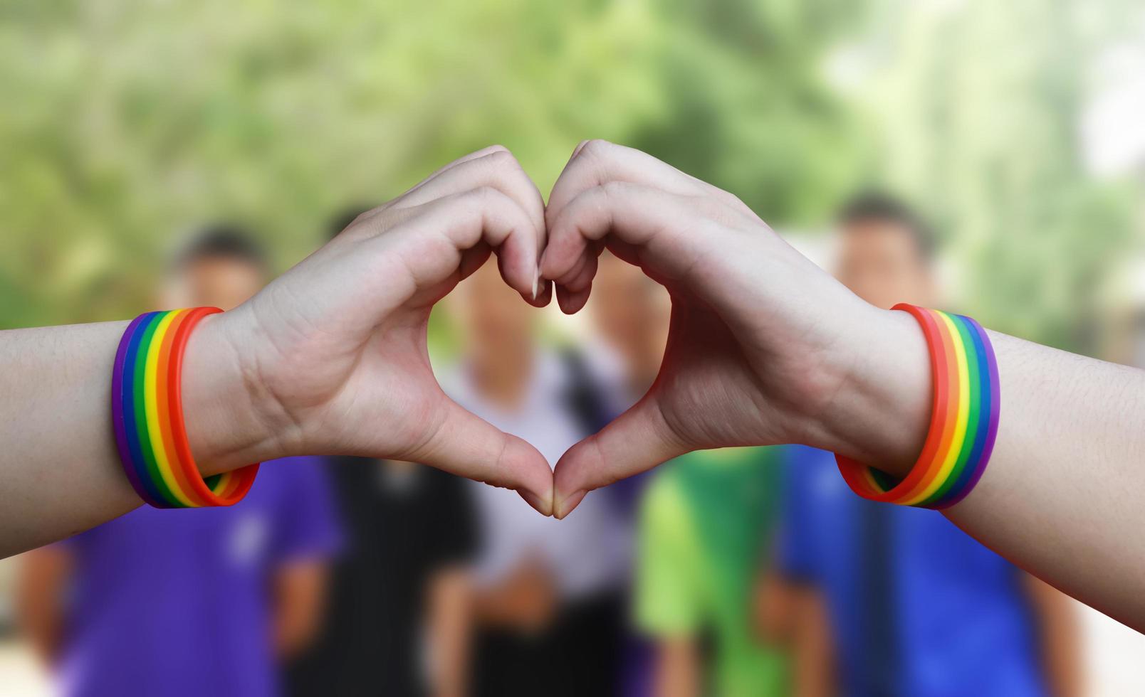 lgbt, dello stesso sesso amore e omosessuale relazioni concetto - vicino su di maschio mani con gay orgoglio arcobaleno consapevolezza braccialetti mostrando cuore gesto con nuvoloso cielo blu sfondo. foto