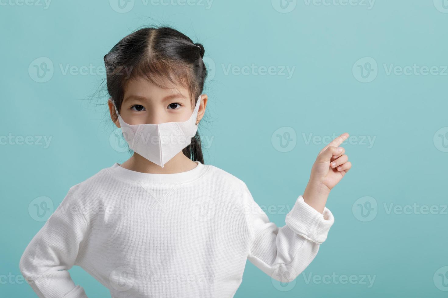 asiatico poco bambino ragazza indossare respiratore maschera per proteggere coronavirus scoppio e puntamento mano per vuoto sfondo, nuovo virus covid-19 a partire dal wuhan Cina concetto, vuoto spazio isolato su blu foto