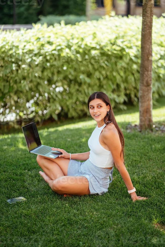 giovane ragazza lavori con un' computer portatile, seduta su il prato. foto