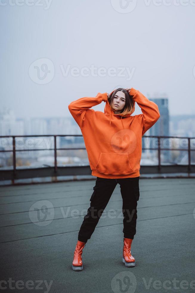 ragazza nel un arancia giacca pose su il tetto di un' edificio nel il città centro foto
