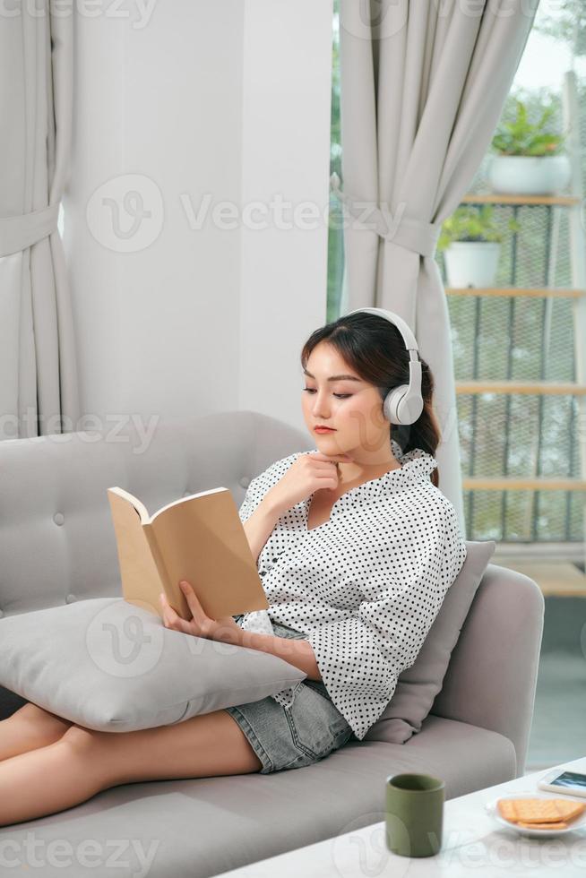 donna con le cuffie che legge un libro in soggiorno seduta sul divano foto