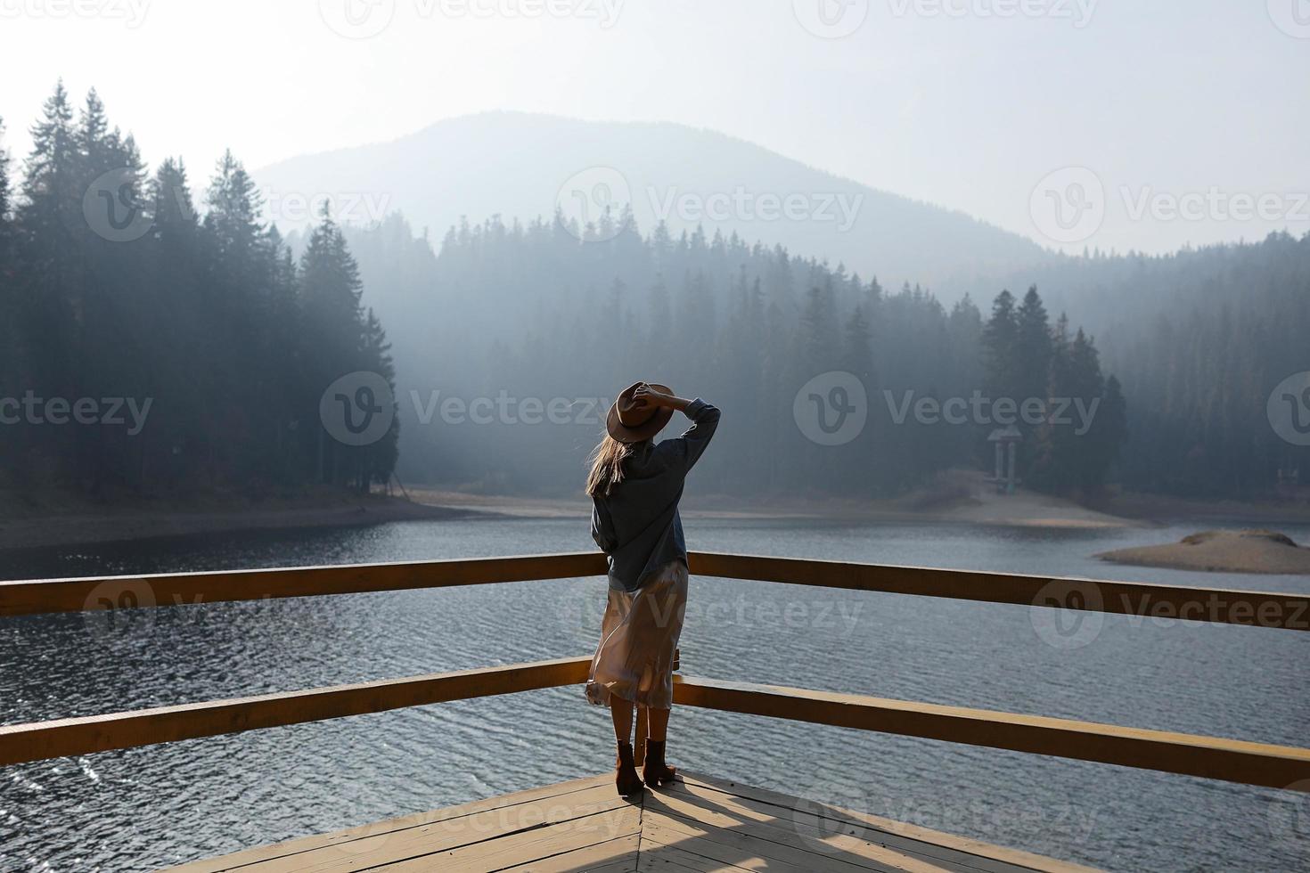 la giovane donna felice in cappello gode della vista del lago in montagne. momenti di relax nella foresta. la vista posteriore della ragazza alla moda gode della freschezza all'aperto. libertà, persone, stile di vita, viaggi e vacanze foto