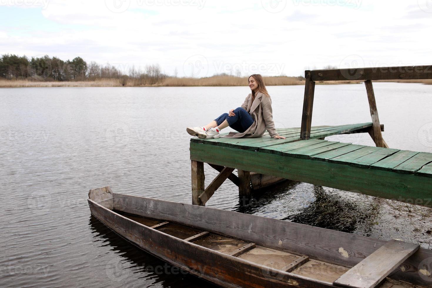 giovane bellissimo donna si siede su il di legno ponte su il fiume a primavera giorno. vecchio barca vicino il costa. selettivo messa a fuoco. foto