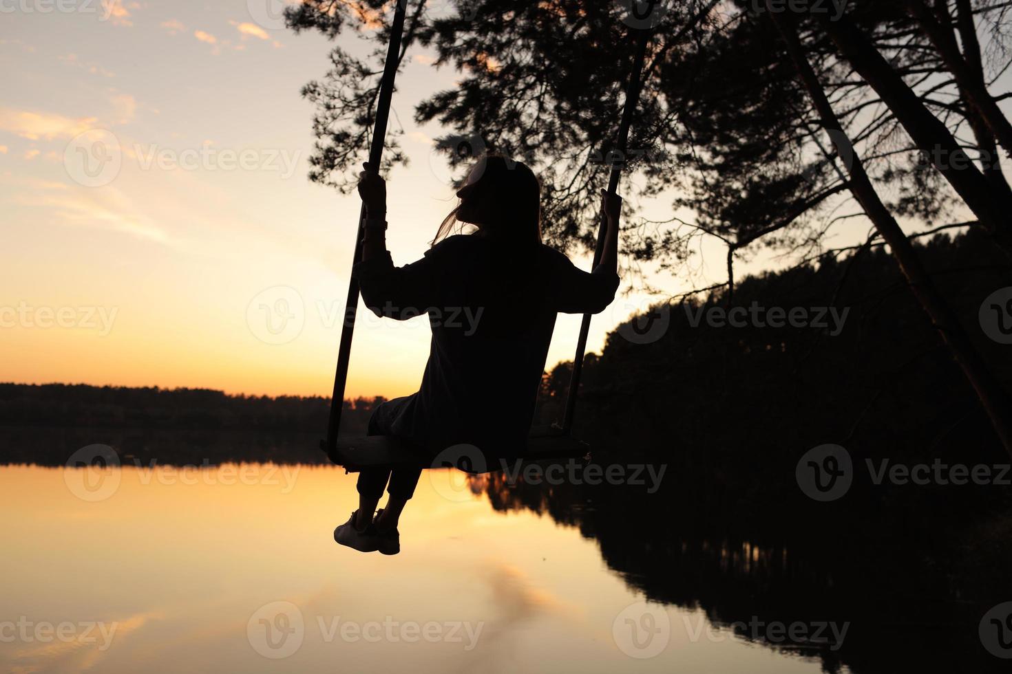 silhouette di un' romantico giovane donna su un' swing al di sopra di lago a tramonto. giovane ragazza viaggiatore seduta su il swing nel bellissimo natura, Visualizza su il lago foto