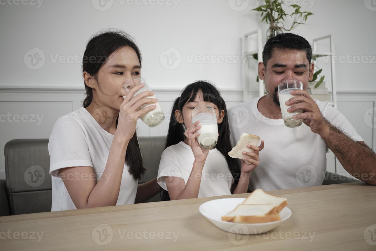 un' salutare asiatico tailandese famiglia, un' poco figlia, e giovane genitori bevanda fresco bianca latte nel bicchiere e pane gioia insieme a un' cenare tavolo nel mattina, benessere nutrizione casa prima colazione pasto stile di vita. foto