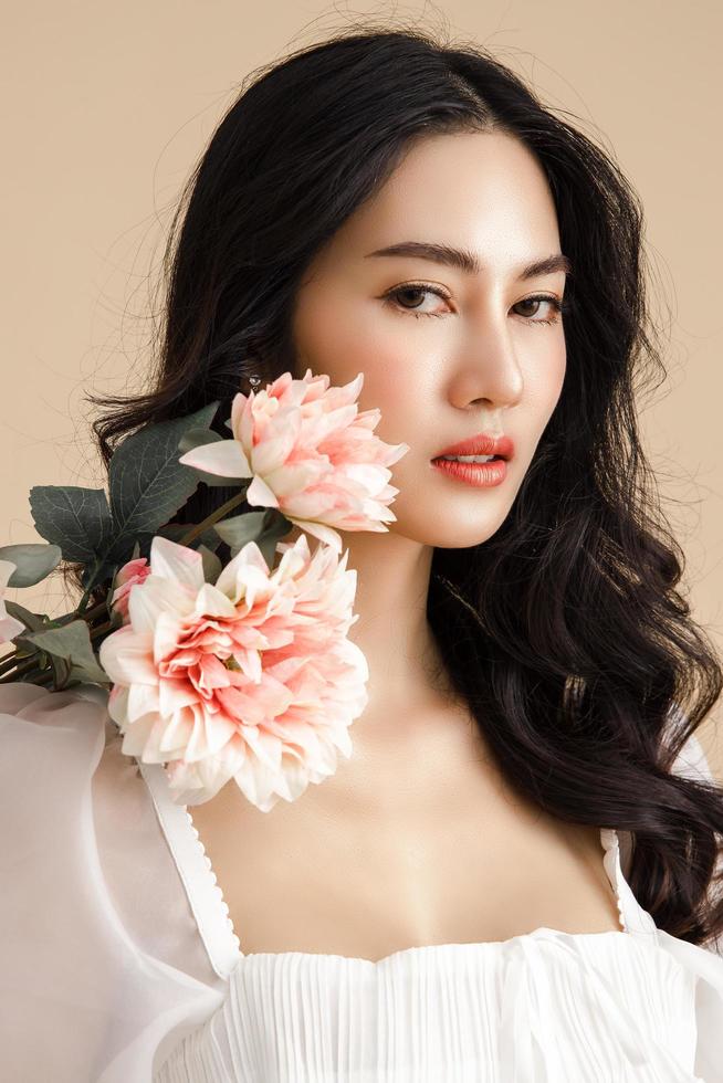 asiatico donna con un' bellissimo viso e Perfetto pulito fresco pelle con fiore. carino femmina modello con naturale trucco e scintillante occhi su beige isolato sfondo. foto