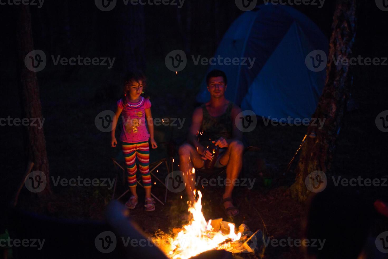 papà e figlia si siedono di notte accanto al fuoco all'aria aperta in estate nella natura. campeggio in famiglia, incontri intorno al fuoco. festa del papà, barbecue. lanterna e tenda da campeggio foto