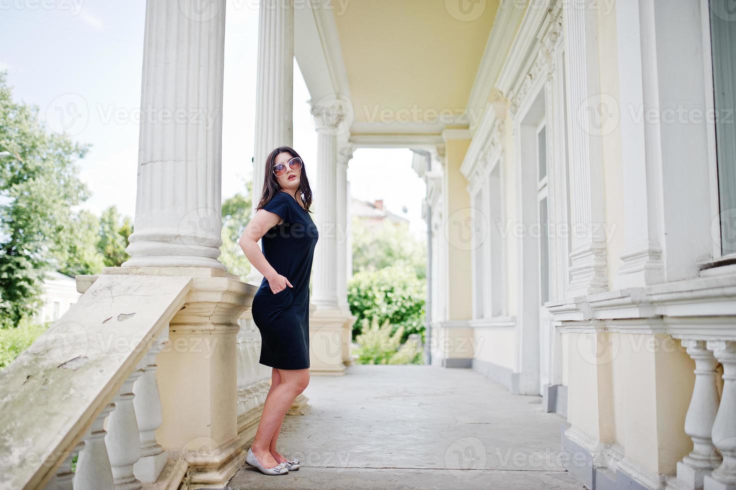 ragazza bruna in abito nero, occhiali da sole in posa contro la vecchia casa d'epoca, in una strada della città. foto