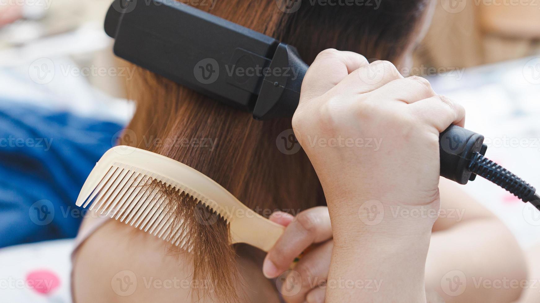 dietro le quinte di una donna dai capelli rossicci che si liscia i capelli con una piastra sul letto della casa. foto