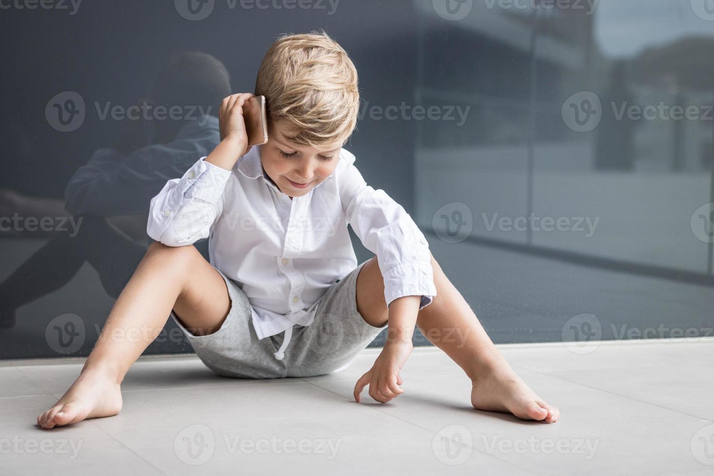 poco ragazzo parlando su mobile Telefono mentre rilassante su il pavimento. foto