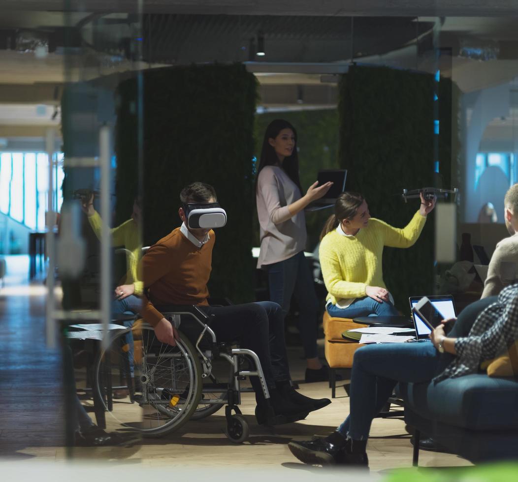 Disabilitato uomo d'affari nel un' sedia a rotelle a opera nel moderno Aperto spazio coworking ufficio con squadra utilizzando virtuale la realtà googles fuco assistenza simulazione foto