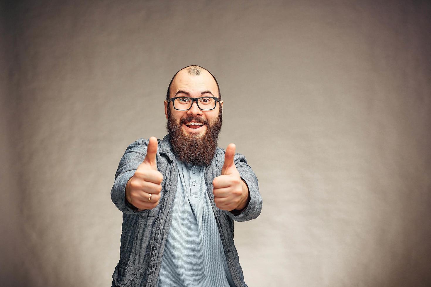 lui stile di vita di un' riuscito giovane uomo con bicchieri , barba, alla moda denim giacca mostrando pollici su, uomini emotivo ritratto nel studio su un' pianura sfondo. foto