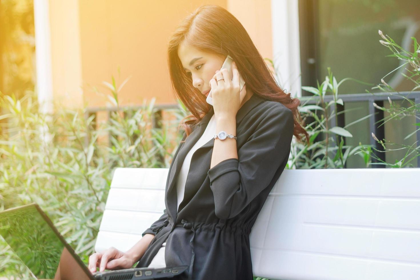 giovane donna asiatica professionale utilizza il suo computer portatile e il telefono su una panchina foto