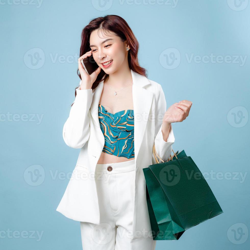 metà corpo di fiducioso bellissimo asiatico attività commerciale donna indossare bianca completo da uomo parlando su mobile Telefono e Tenere shopping Borsa su isolato blu sfondo. vuoto schermo smartphone concetto. foto