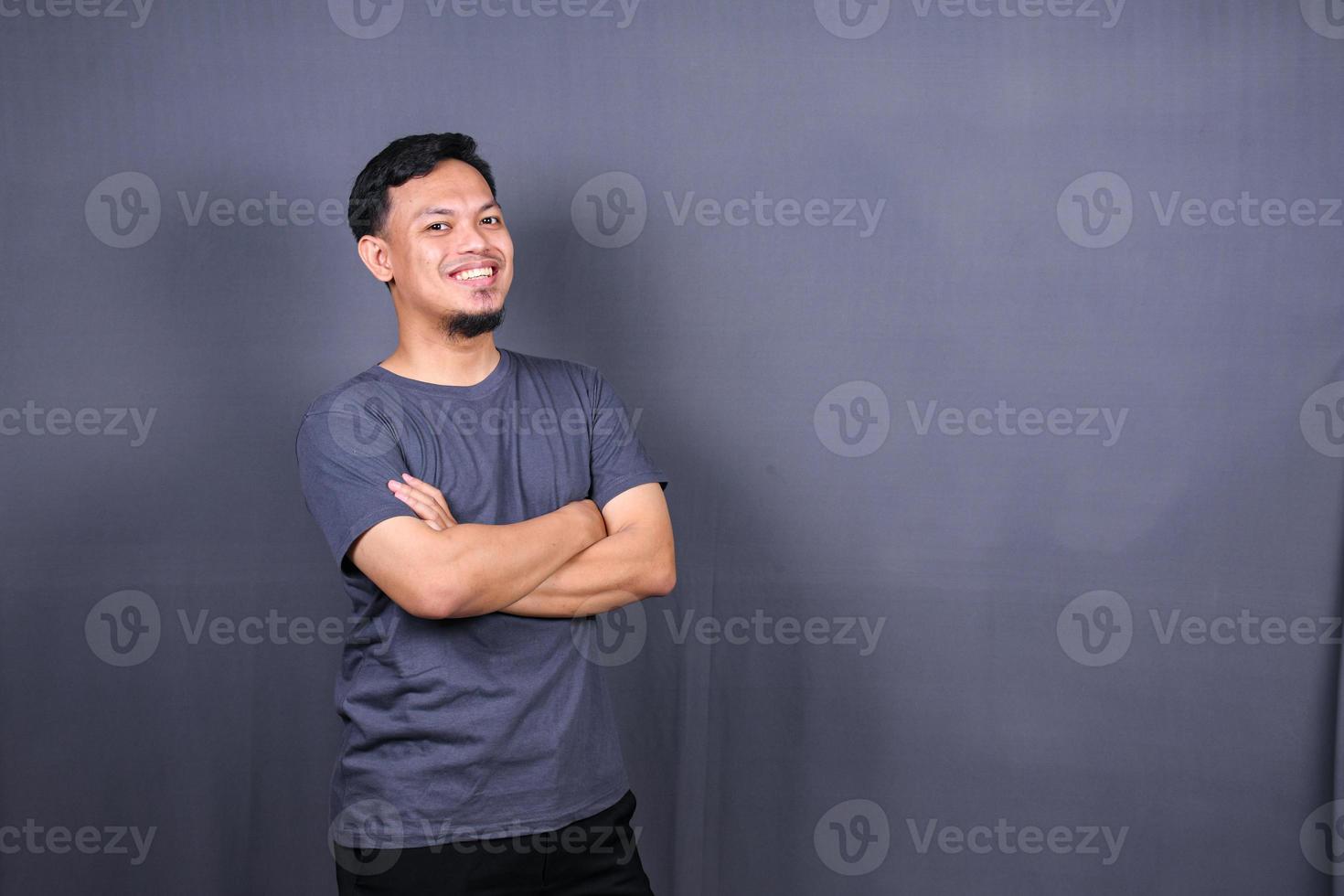 sorridente bello uomo nel grigio maglietta in piedi con attraversato braccia isolato su grigio sfondo foto