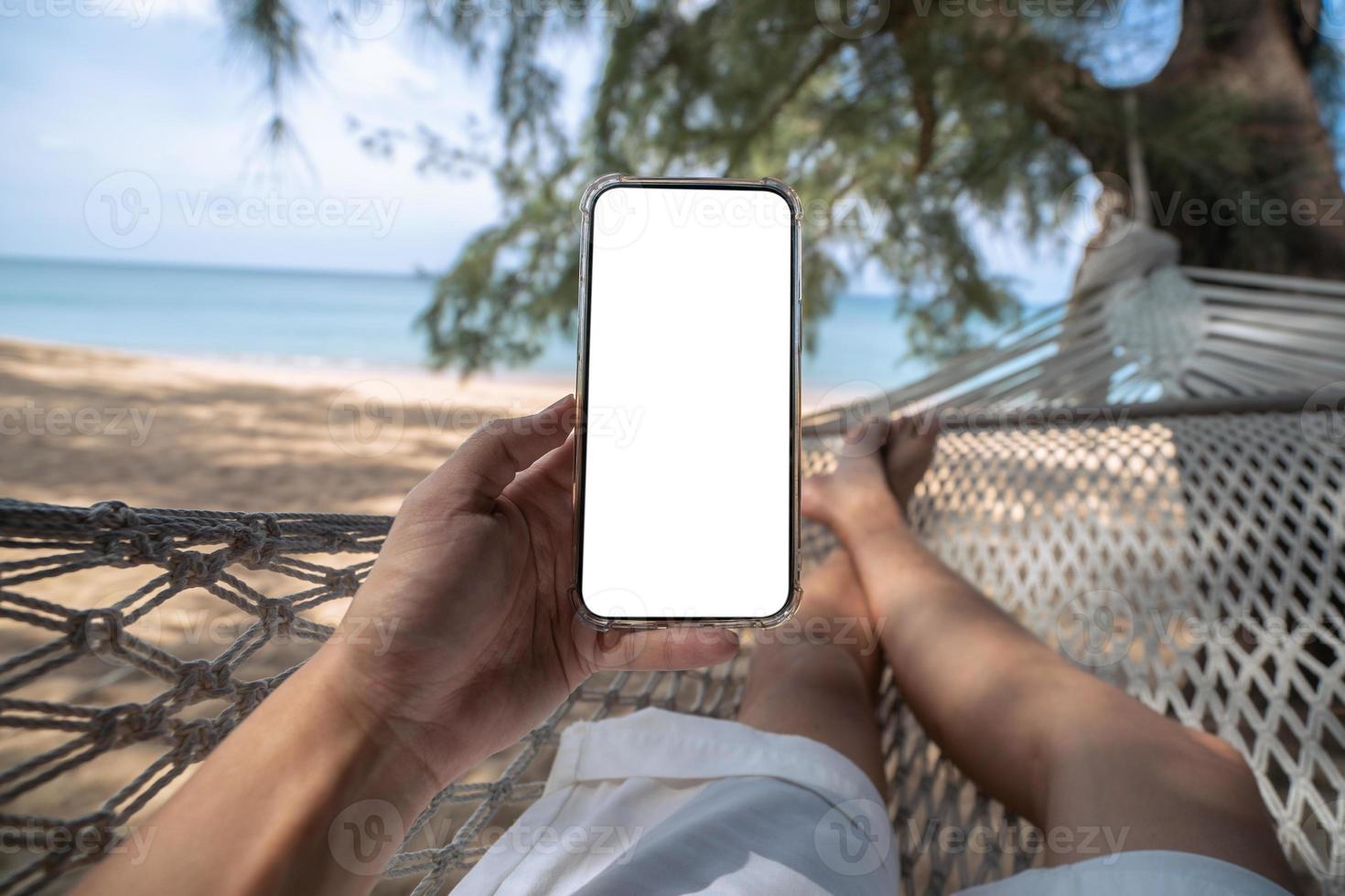 mano Tenere finto su mobile con bianca scena mentre posa su amaca swing fra alberi su il spiaggia. foto