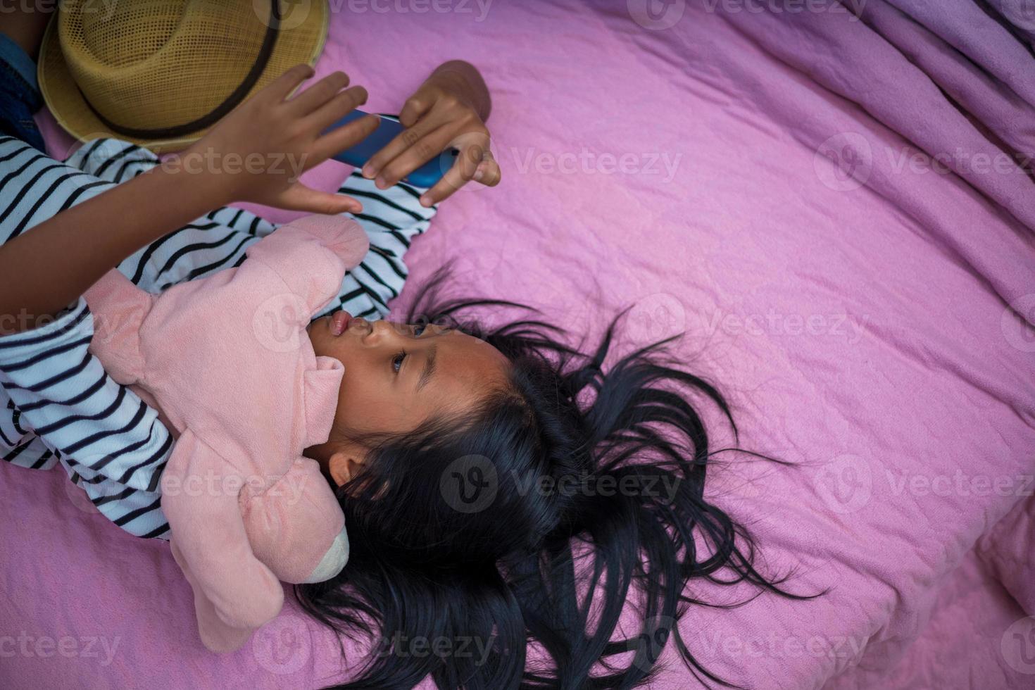 asiatico ragazza Guardando soddisfare su sua letto nel il mattina utilizzando un' smartphone foto