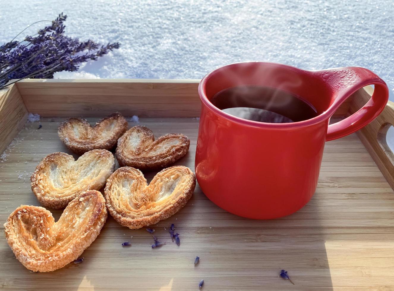 rosso boccale con biscotti su il rosso neve ceramica tazza con caldo tè e soffio Pasticcino zucchero biscotti nel il forma di cuori su un' di legno vassoio, su neve su un' soleggiato giorno, su di messa a fuoco sfondo, concetto di amore foto