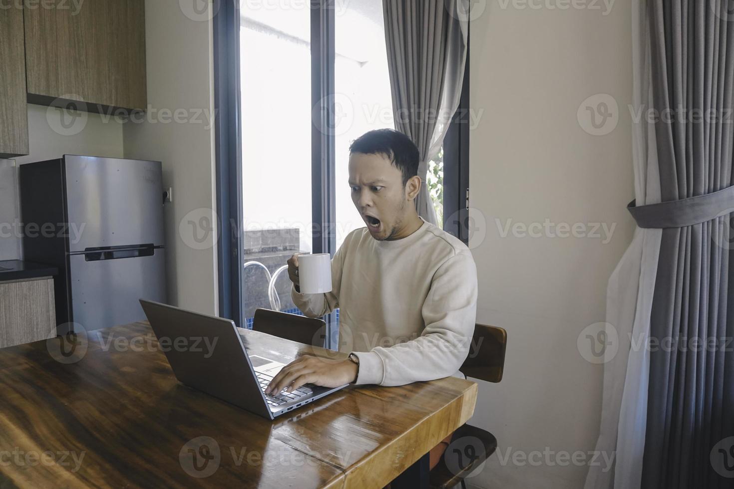 un' giovane asiatico uomo spende il suo tempo a casa seduta nel il cenare camera scioccato mentre Lavorando su il suo il computer portatile mentre Tenere un' boccale foto