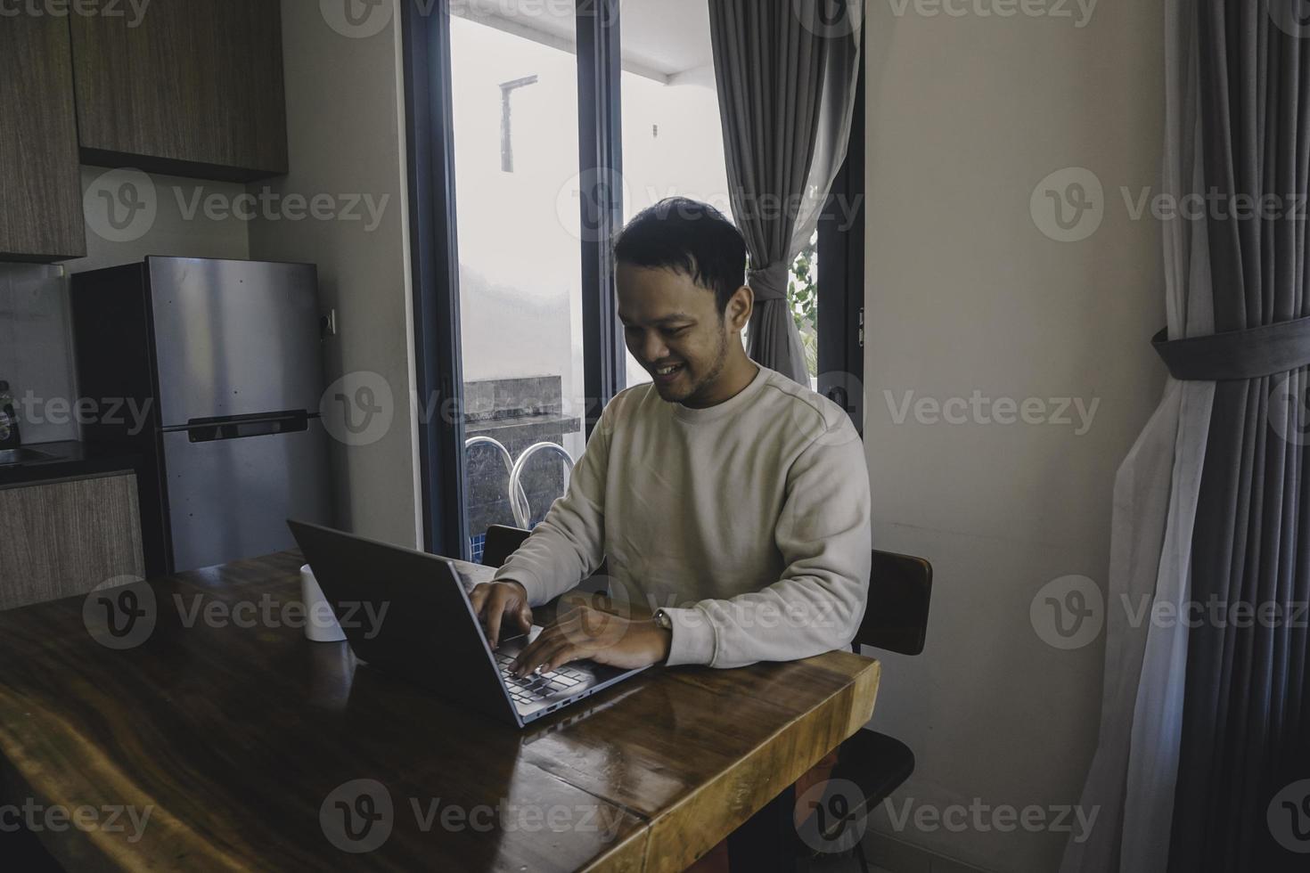 un' giovane asiatico uomo spende il suo tempo a casa seduta nel il cenare camera felicemente Lavorando su il suo il computer portatile foto