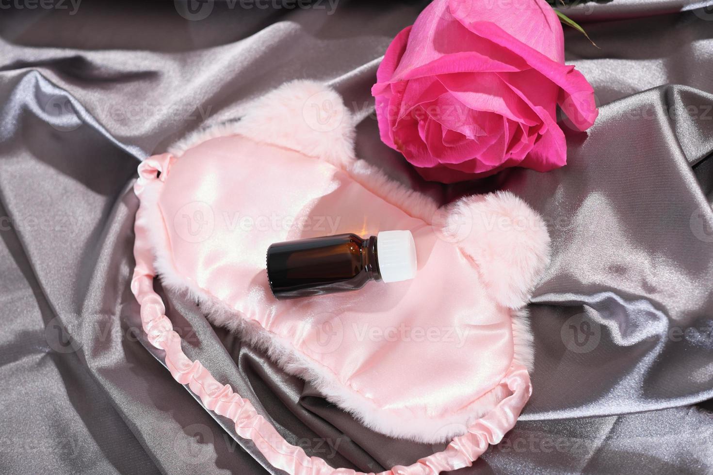 essenziale rosa petalo olio nel bicchiere ambra bottiglia. addormentato maschera e rosa fiore su seta raso letto fogli. bene dormire, aromaterapia concetto, naturale rimedio per trattamento di depressione, insonnia fatica foto