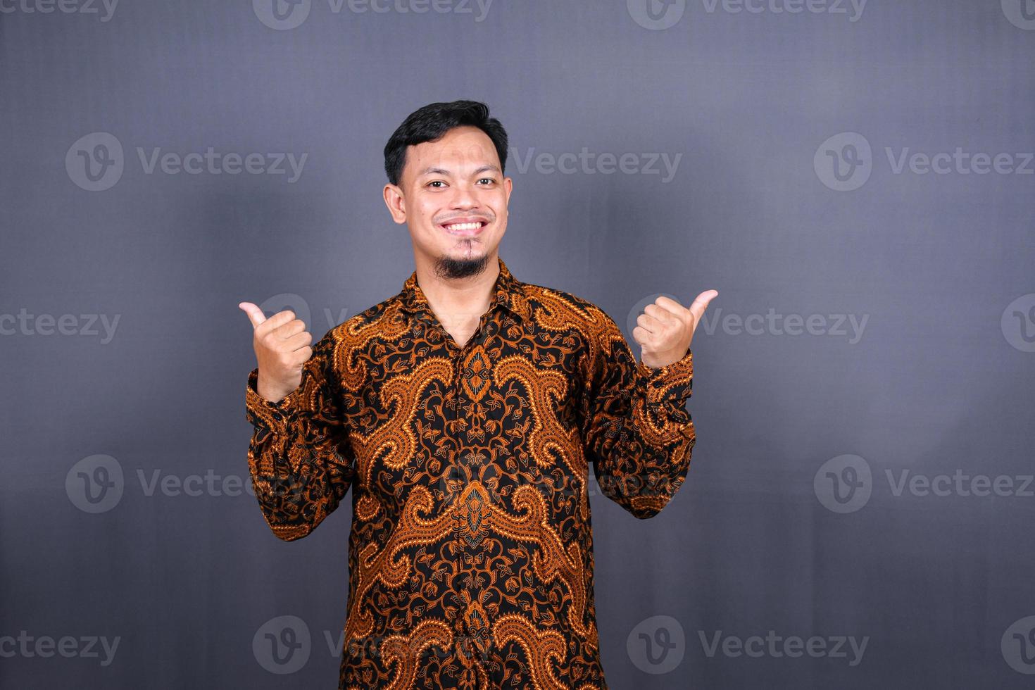 ritratto di un' contento giovane uomo nel batik Indonesia puntamento dita lontano a copia spazio isolato al di sopra di grigio sfondo foto