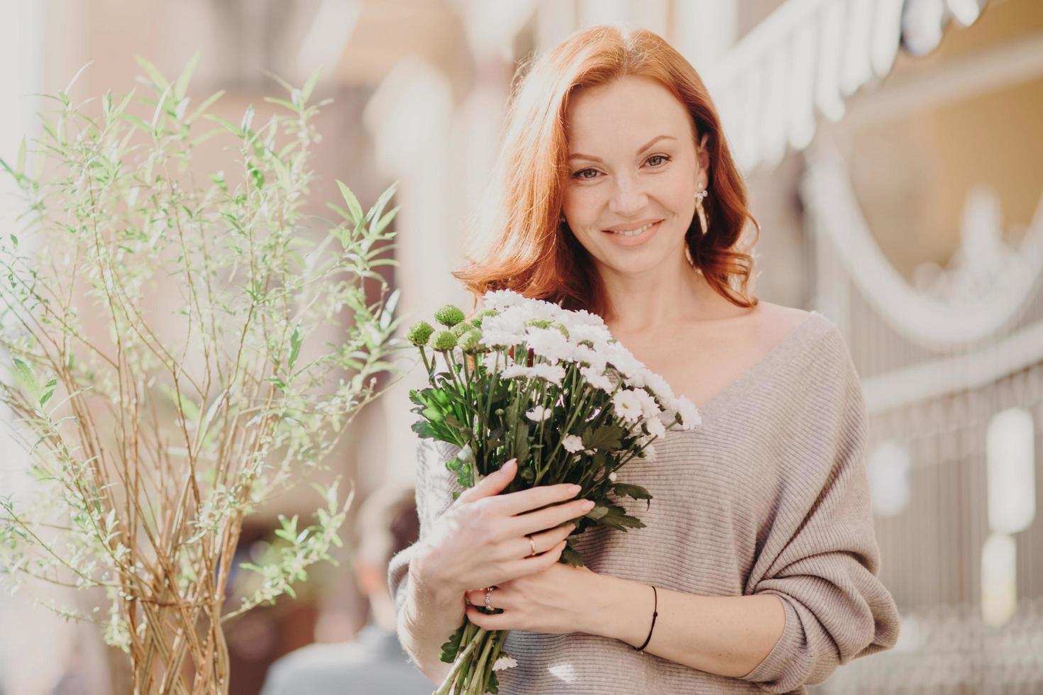 scatto all'aperto di una giovane femmina di zenzero dall'aspetto piacevole con un'espressione soddisfatta, indossa un maglione marrone, tiene un mazzo di fiori bianchi, cammina per strada su sfondo sfocato. soleggiata giornata di primavera foto