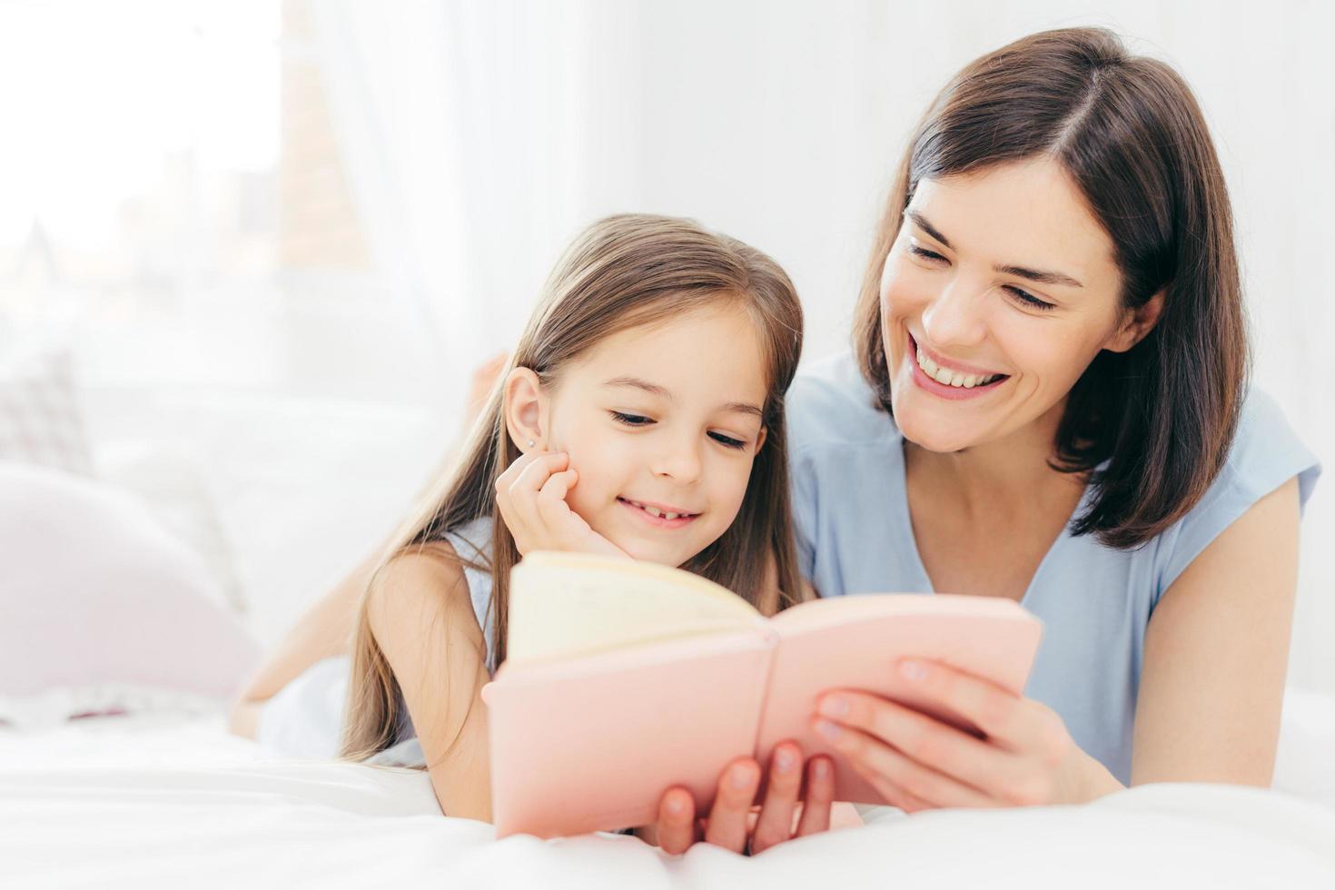 la bella madre e la sua piccola figlia leggono un libro interessante in camera da letto, si sdraiano su un letto comodo durante la mattina. mamma adorabile legge fiabe per la sua bambina, che è molto curiosa foto