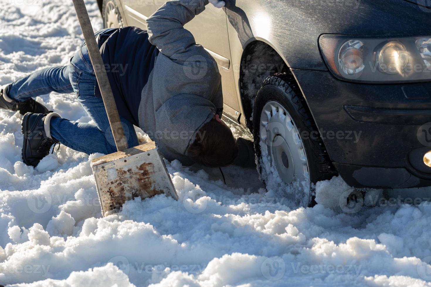 uomo Lavorando a auto incollato nel neve su ginocchio con pala a luce del giorno fuori strada foto