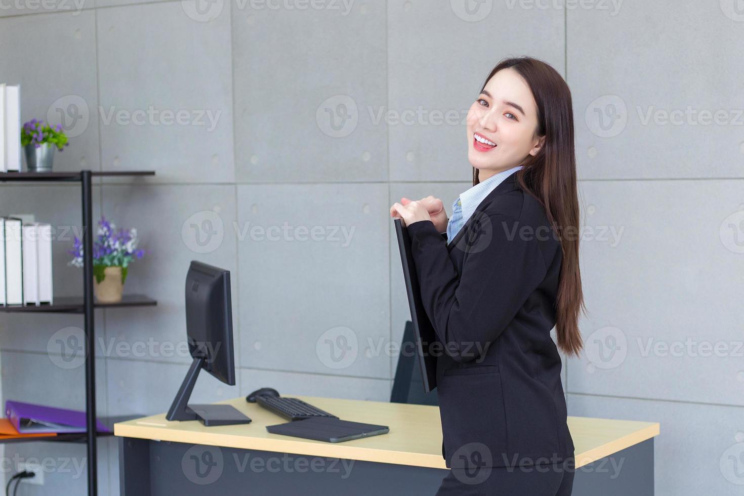 asiatico professionale attività commerciale giovane donna nel nero completo da uomo sorrisi felicemente In piedi con fiducia e Guarda a il telecamera mentre lei lavori e detiene appunti nel ufficio. foto