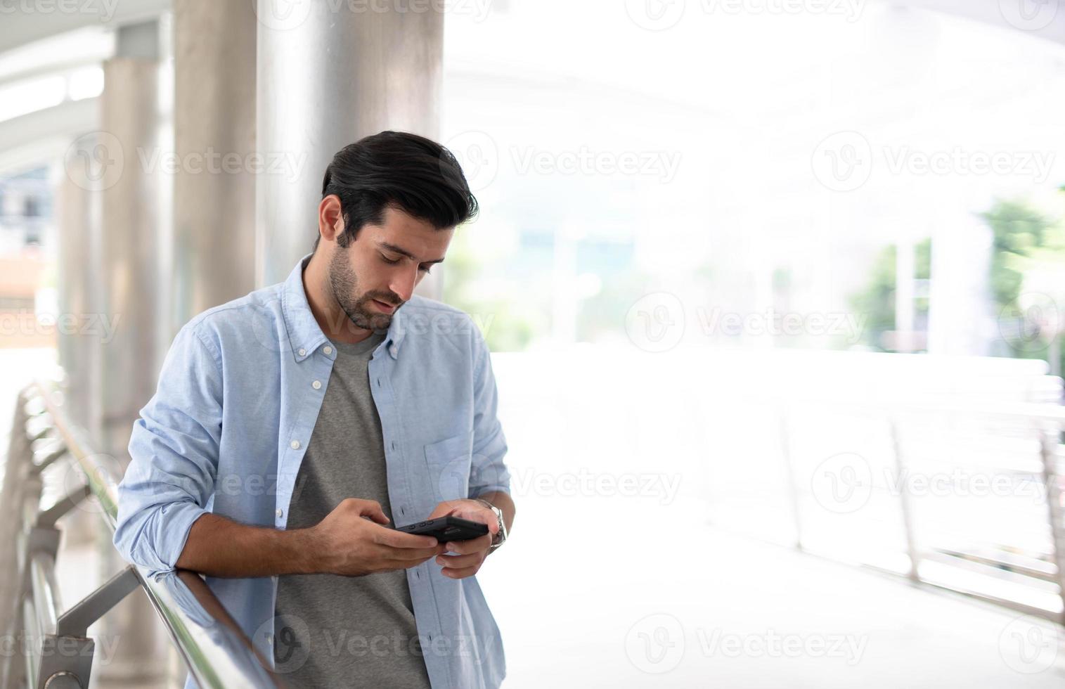giovane caucasico uomo utilizzando inteligente Telefono e sorridente mentre in attesa il suo amico a il fuori. il uomo Tenere e utilizzando cellulare per ricerca dati e sociale media su Internet. foto