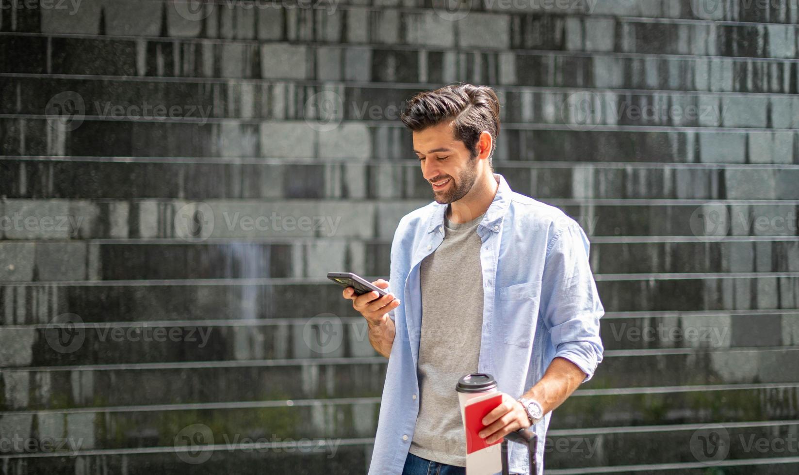 giovane caucasico uomo utilizzando smartphone Tenere il caffè tazza e traino il valigia in attesa il suo amico per viaggio insieme. foto