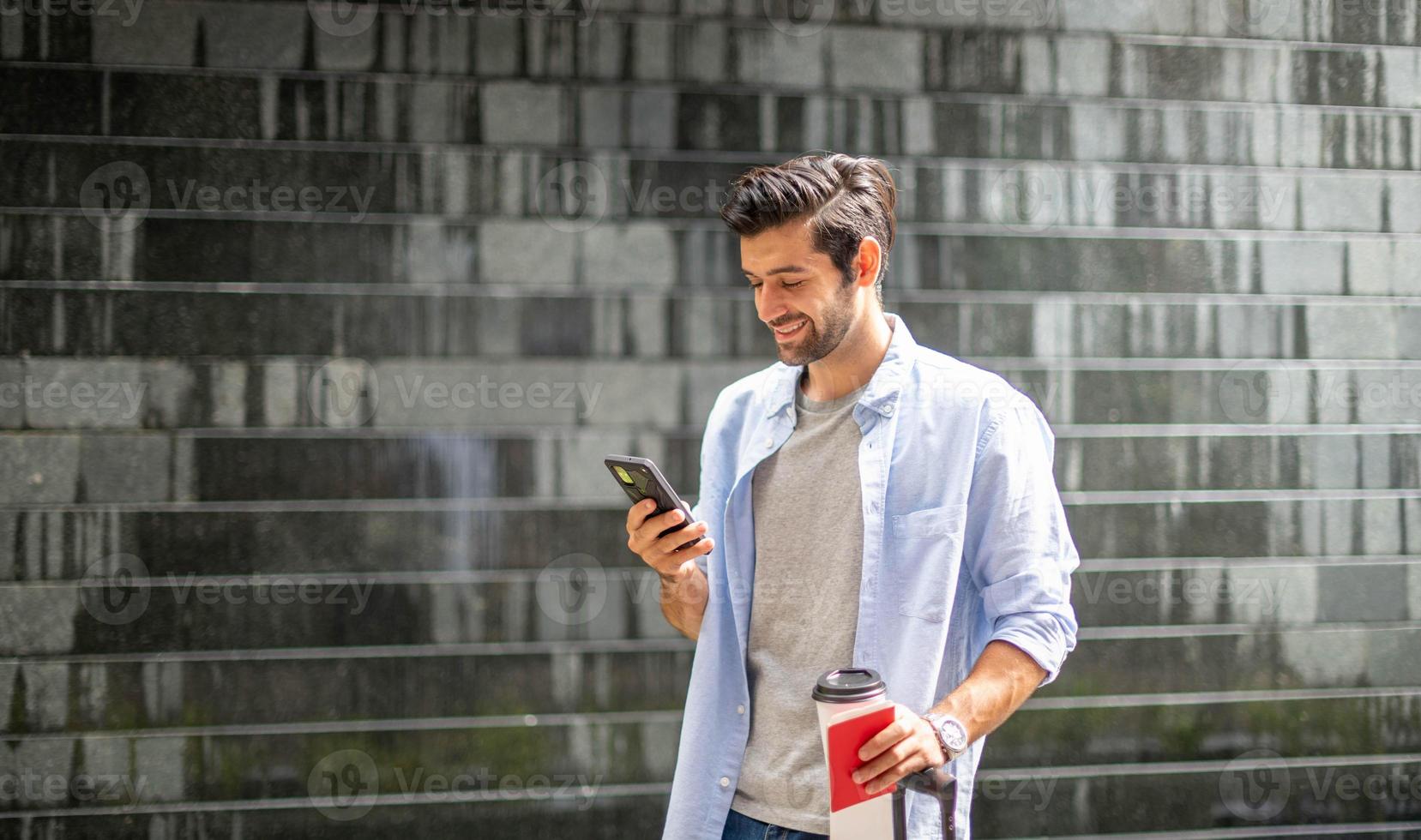 giovane caucasico uomo utilizzando smartphone Tenere il caffè tazza e traino il valigia in attesa il suo amico per viaggio insieme. foto