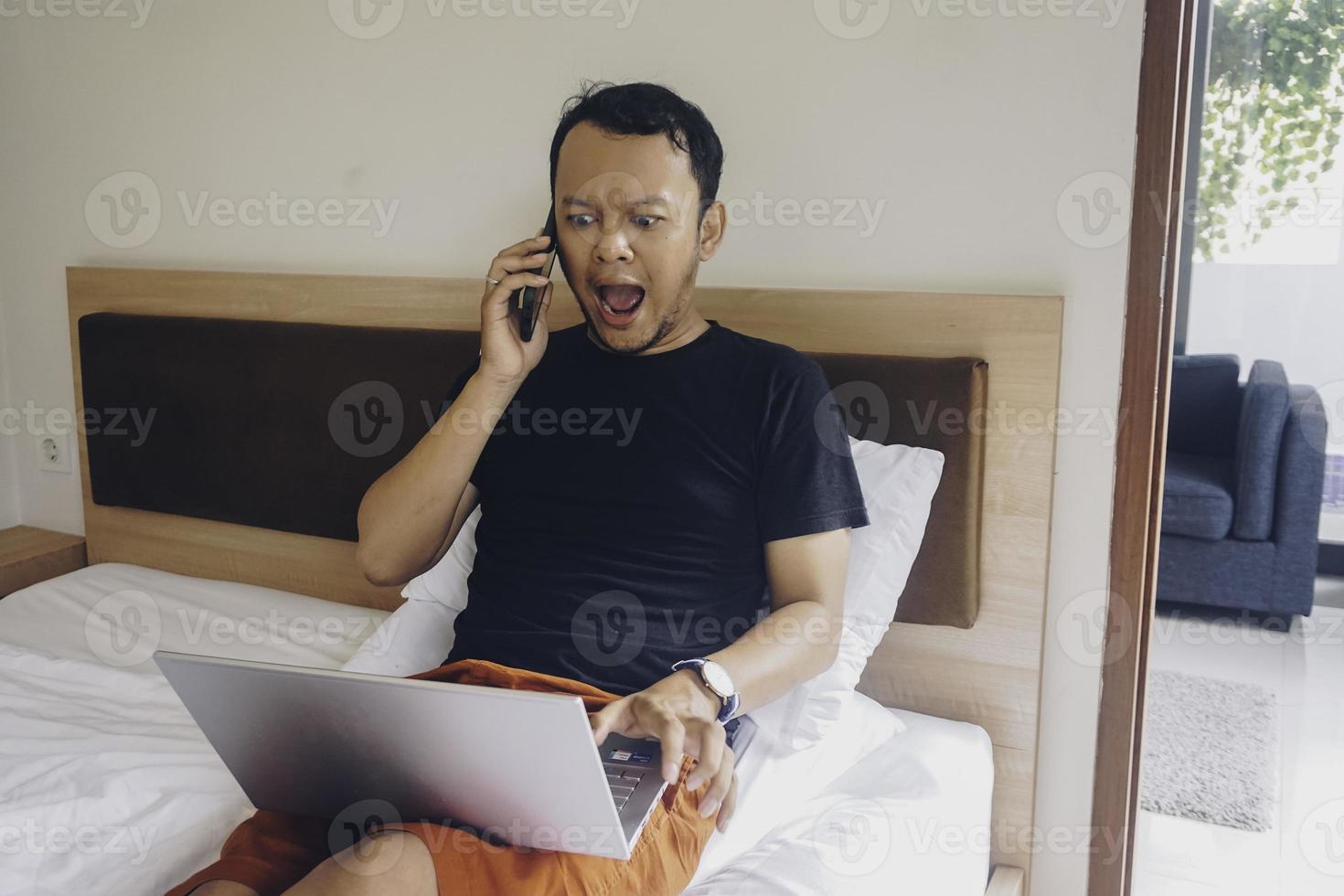 contento giovane uomo Lavorando a partire dal il letto. hold il il computer portatile su il ginocchia e parlando su il Telefono. foto