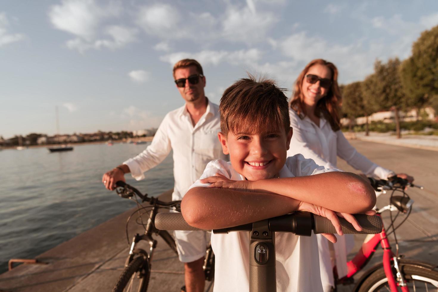 contento famiglia godendo un' bellissimo mattina di il mare insieme, genitori equitazione un' bicicletta e loro figlio equitazione un elettrico scooter. selettivo messa a fuoco foto