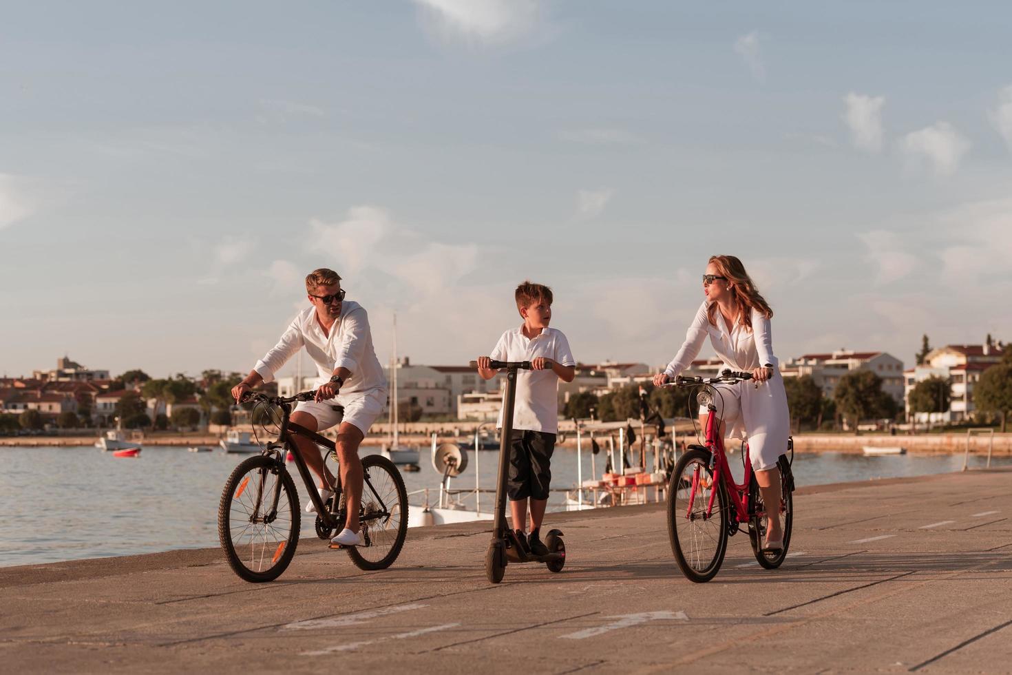 contento famiglia godendo un' bellissimo mattina di il mare insieme, genitori equitazione un' bicicletta e loro figlio equitazione un elettrico scooter. selettivo messa a fuoco foto