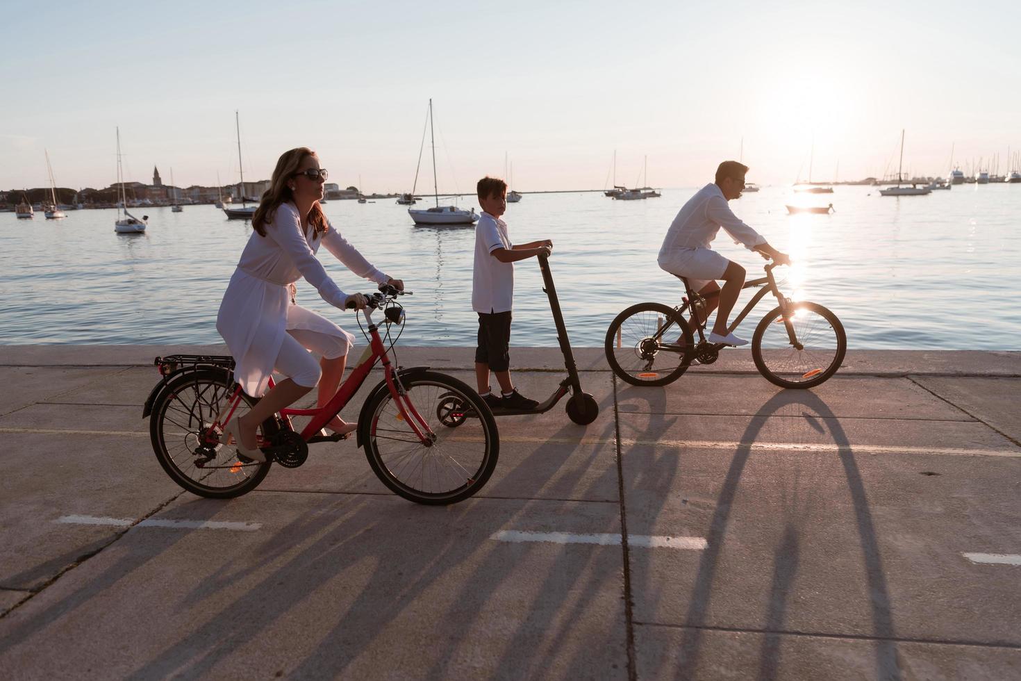 contento famiglia godendo un' bellissimo mattina di il mare insieme, genitori equitazione un' bicicletta e loro figlio equitazione un elettrico scooter. selettivo messa a fuoco foto
