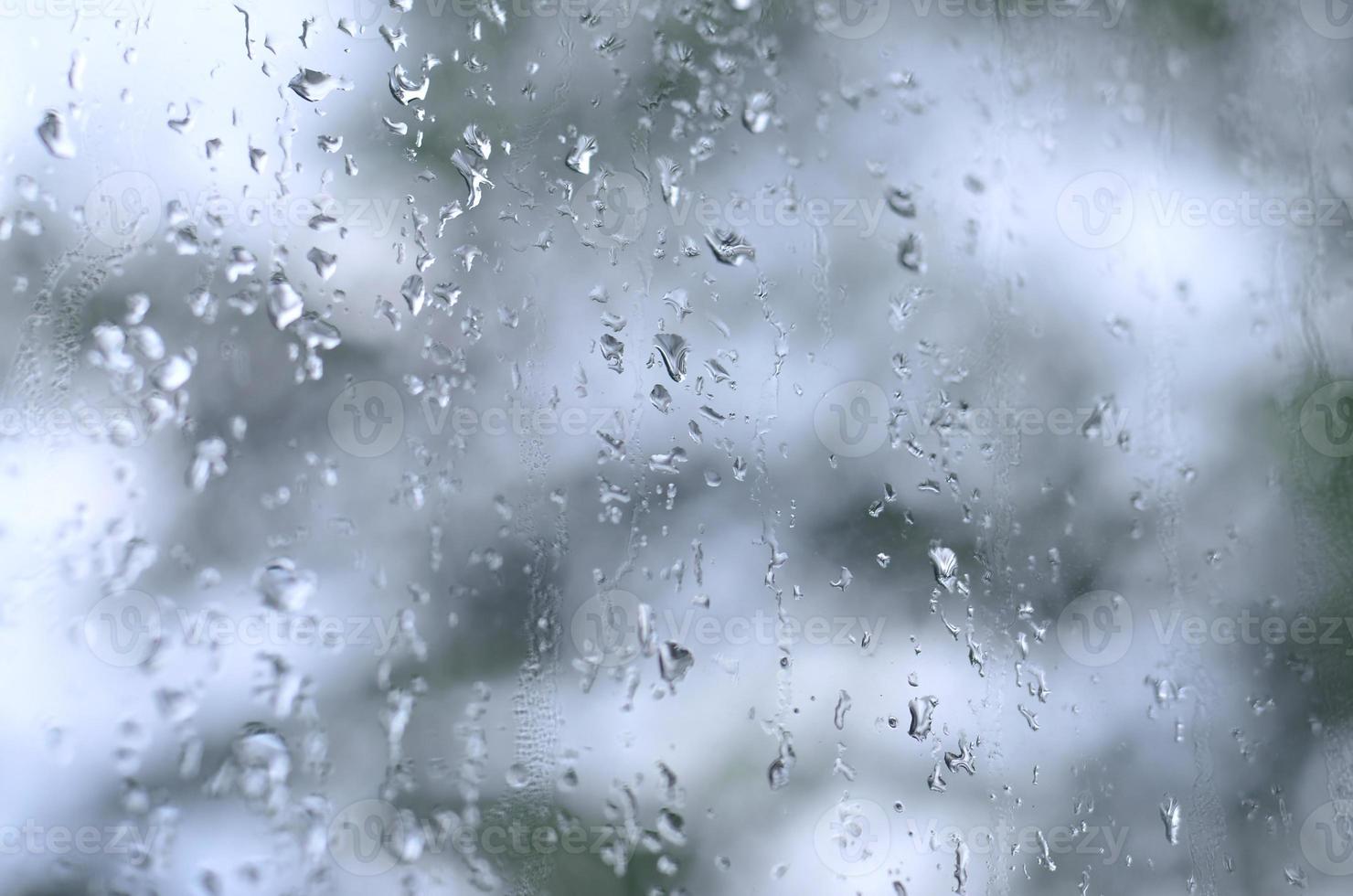 un' foto di pioggia gocce su il finestra bicchiere con un' sfocato Visualizza di il fioritura verde alberi. astratto Immagine mostrando nuvoloso e piovoso tempo metereologico condizioni