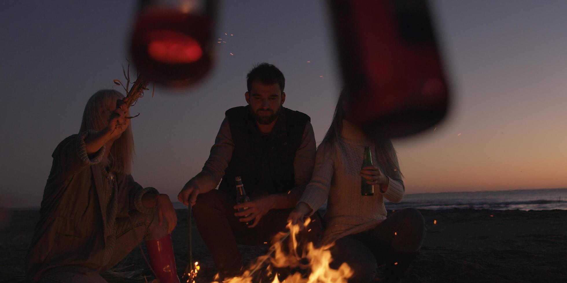 giovane amici fabbricazione un' crostini con birra in giro fuoco di bivacco a spiaggia foto