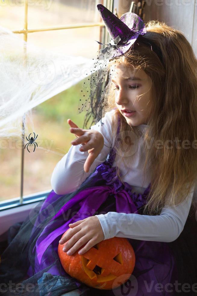 una ragazza in costume da strega sul davanzale vicino alla finestra sta giocando con una ragnatela e un ragno, decorando la casa per le vacanze di Halloween. scenario terribile, paura e orrore, zucche foto