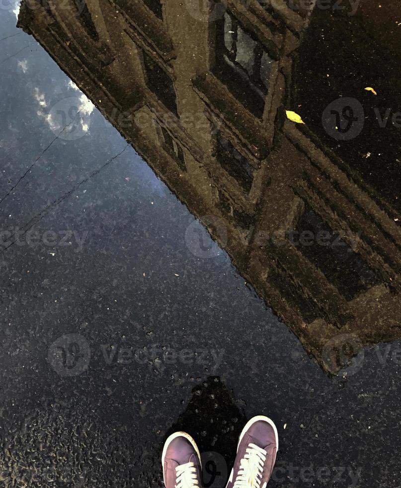 riflessione nel un' pozzanghera di un' edificio e il gambe di un' uomo nel scarpe da ginnastica. foto