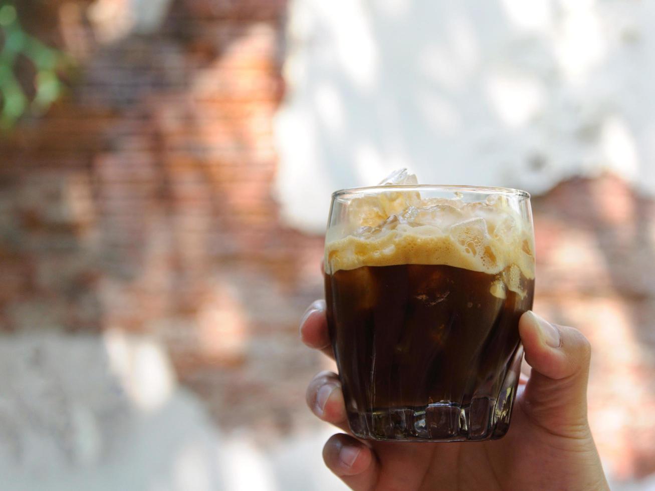 il mano Tenere un' bicchiere di scosso caffè espresso miscelazione ghiaccio cubo con caffè per freddo bevanda per godere caldo tempo metereologico di il estate con il sfondo di vecchio mattone parete foto