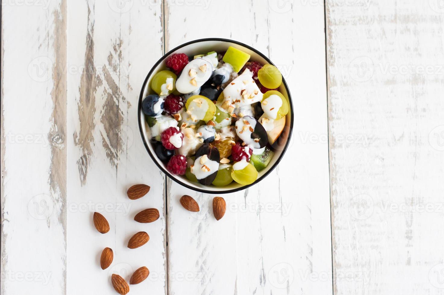 andiamo avere un' fresco frutta insalata con Yogurt e mandorle nel un' ciotola su un' bianca di legno tavolo. salutare mangiare. superiore Visualizza. foto