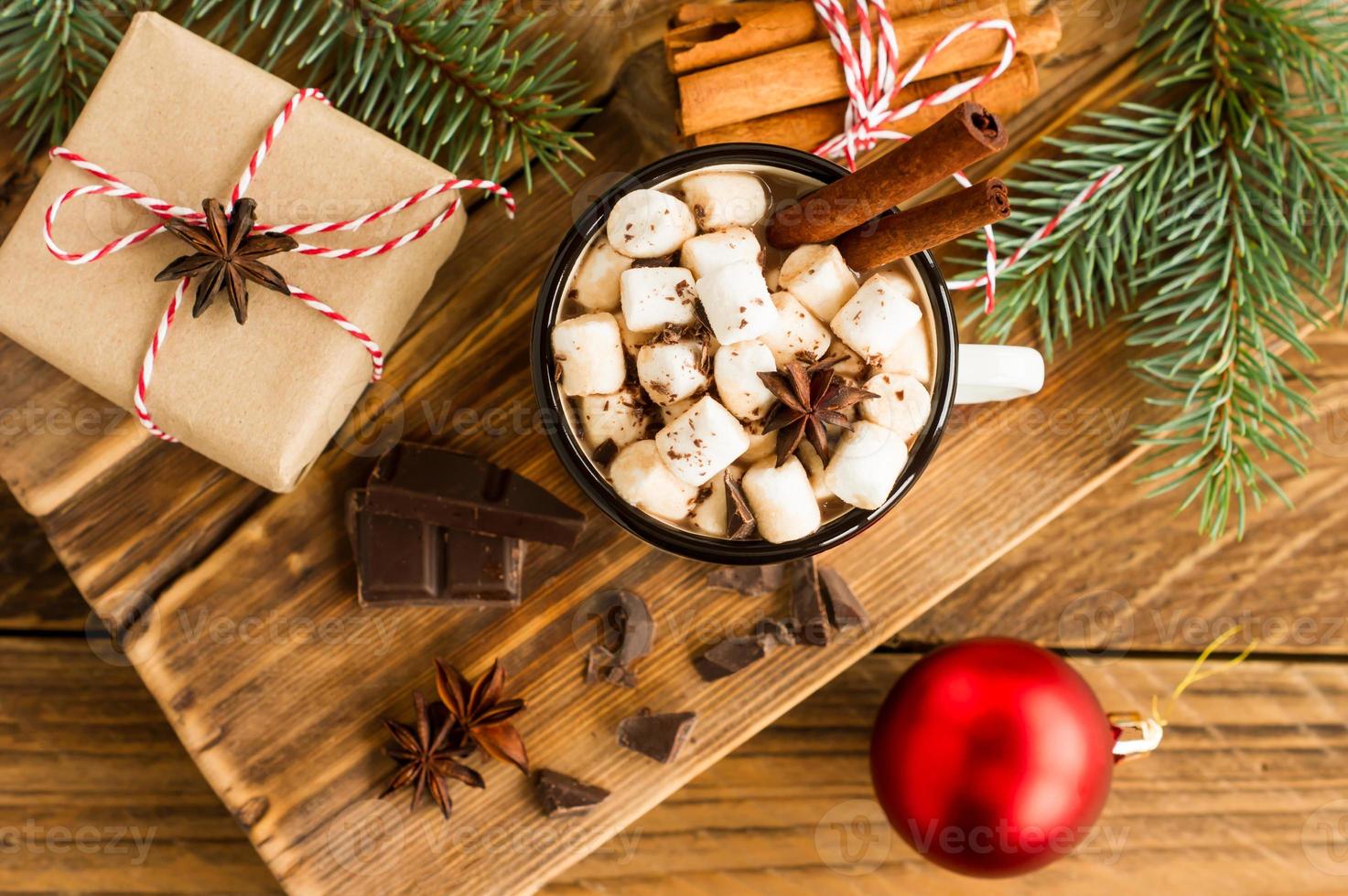 smaltato boccale con cioccolato bevanda e marshmallow su un' di legno nazione tavolo con un' regalo scatola e pezzi di cioccolato qui vicino. foto