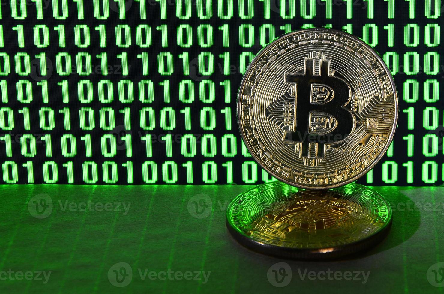 un' paio di bitcoin bugie su un' cartone superficie su il sfondo di un' tenere sotto controllo raffigurante un' binario codice di luminosa verde zeri e uno unità su un' nero sfondo. Basso chiave illuminazione foto