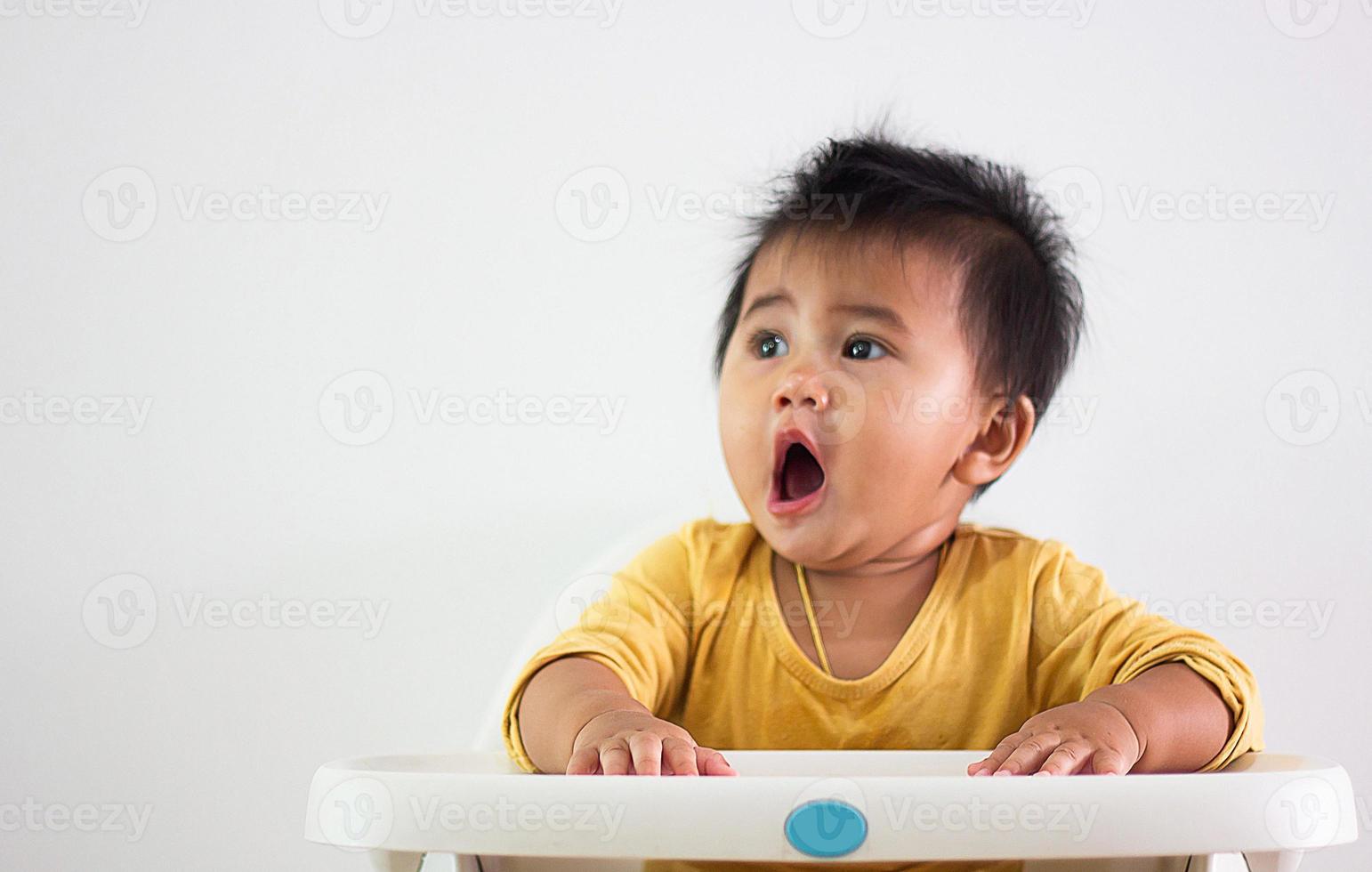 bambino ragazza di asiatico etnia giallo camicia si siede pazientemente nel un' sedia solo a casa. espressione di emozione, eccitazione, infanzia, e infantilismo. su quello carino Wow viso felicità vita foto