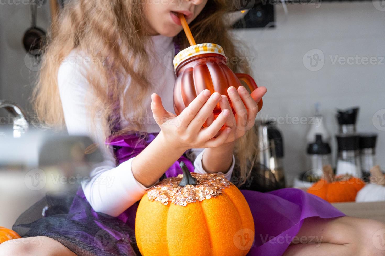 bambino bevande zucca succo, verdura frullato o cocktail a partire dal un' boccale con un' cannuccia. cucina decora per Halloween. ragazza nel un' strega costume. autunno accogliente nel casa foto