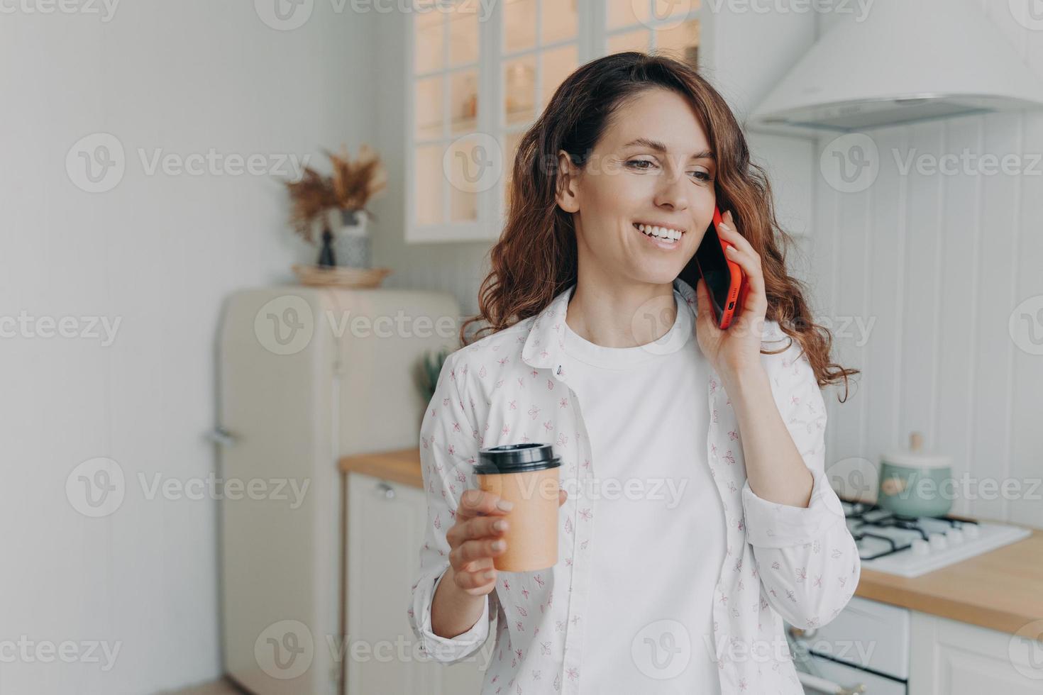 sorridente femmina Tenere porta via caffè, parlando su Telefono, godendo piacevole conversazione nel cucina foto