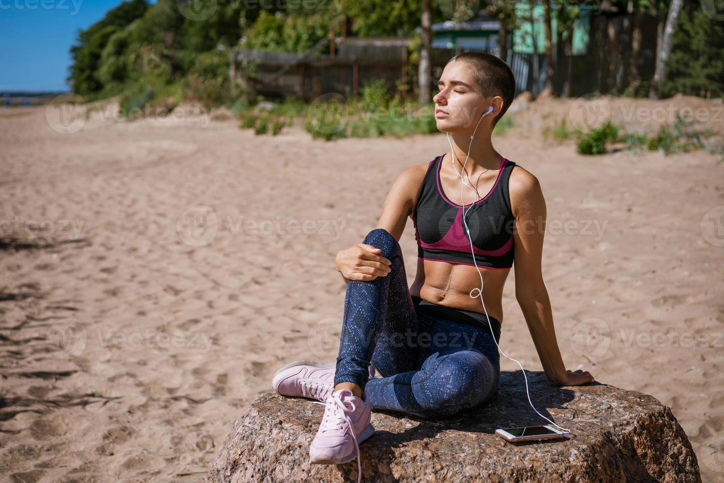 giovane donna vestito nel abbigliamento sportivo si siede su spiaggia per apprendimento musica ascoltando foto