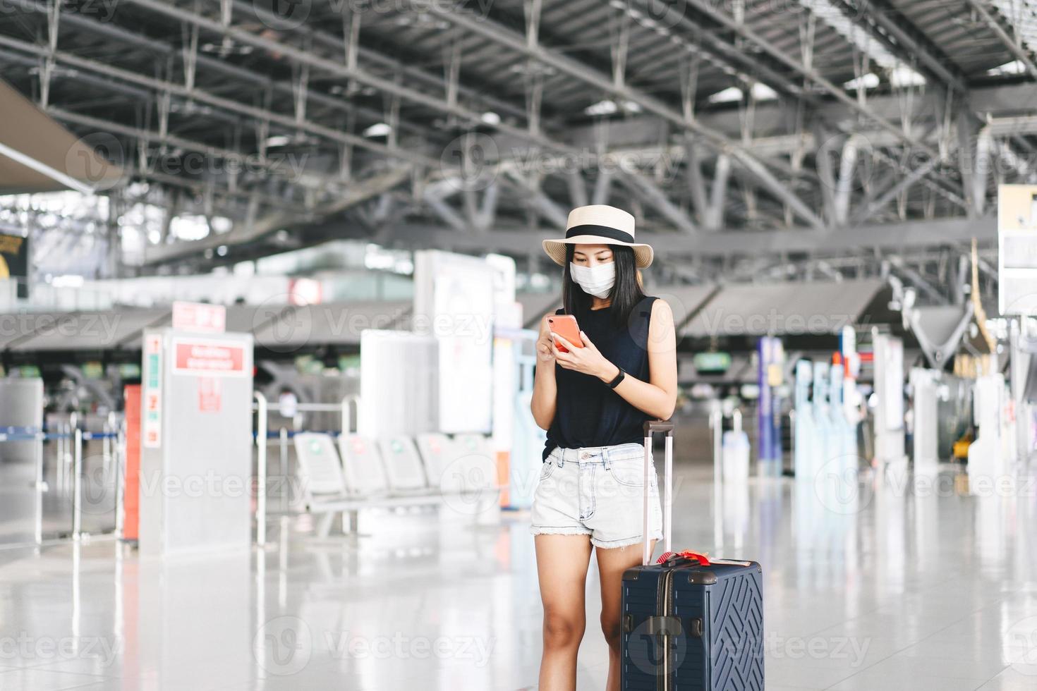 adulto turista donna con bagaglio indossare maschera per covid-19 dai un'occhiata nel con mobile Telefono a aeroporto terminale. foto